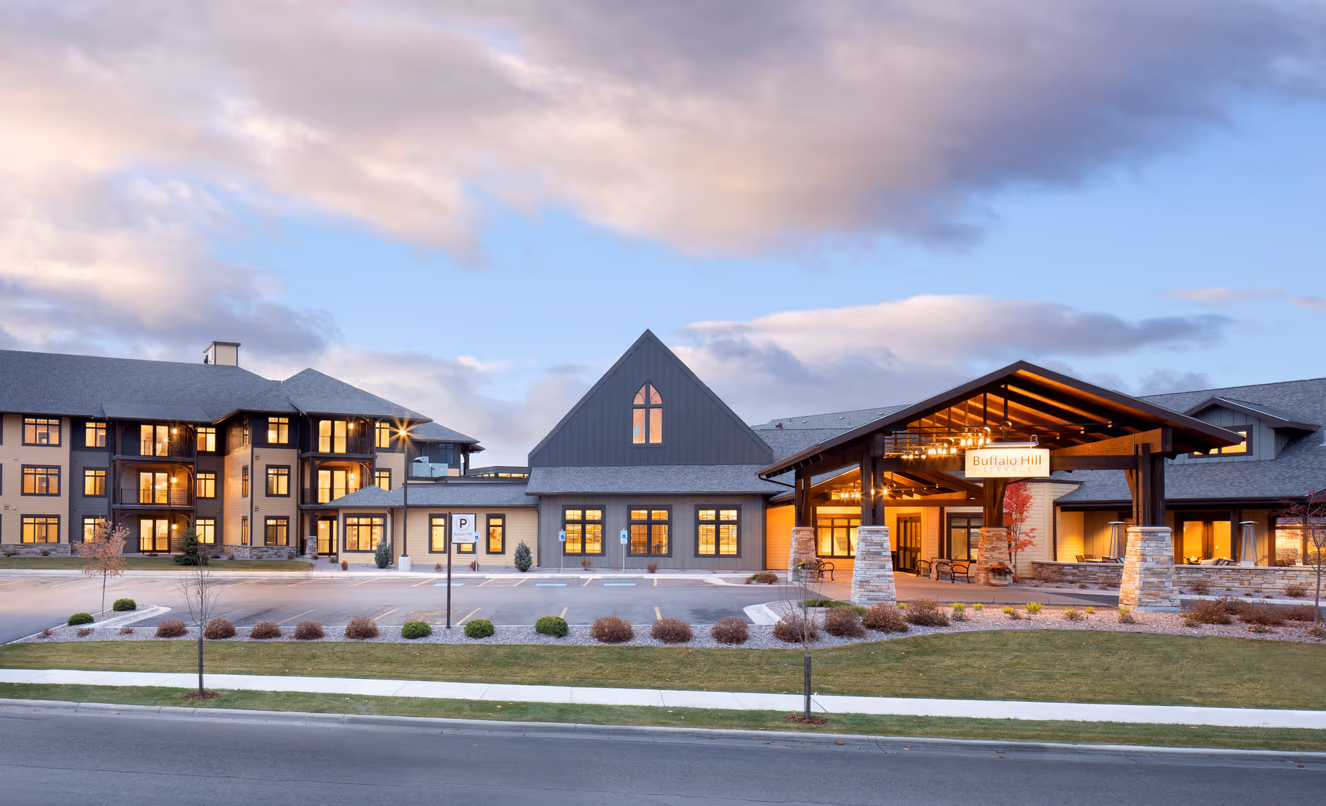 Exterior view of a senior living facility named Immanuel Living during dusk, showing a large building with multiple windows lit from inside, a covered entrance with a sign that reads 'Buffalo Hill', landscaped grounds, and a parking area in front.
