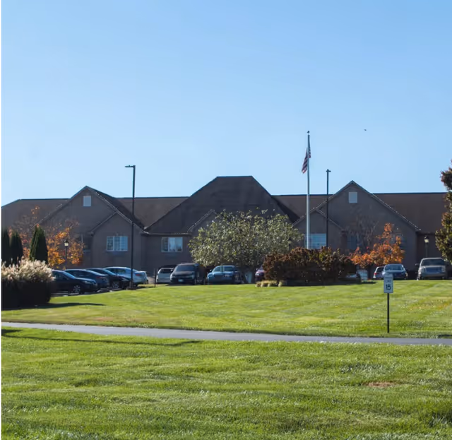 Exterior view of a senior living facility building with a large green lawn in the foreground, several parked cars, trees with autumn foliage, and an American flag on a flagpole.