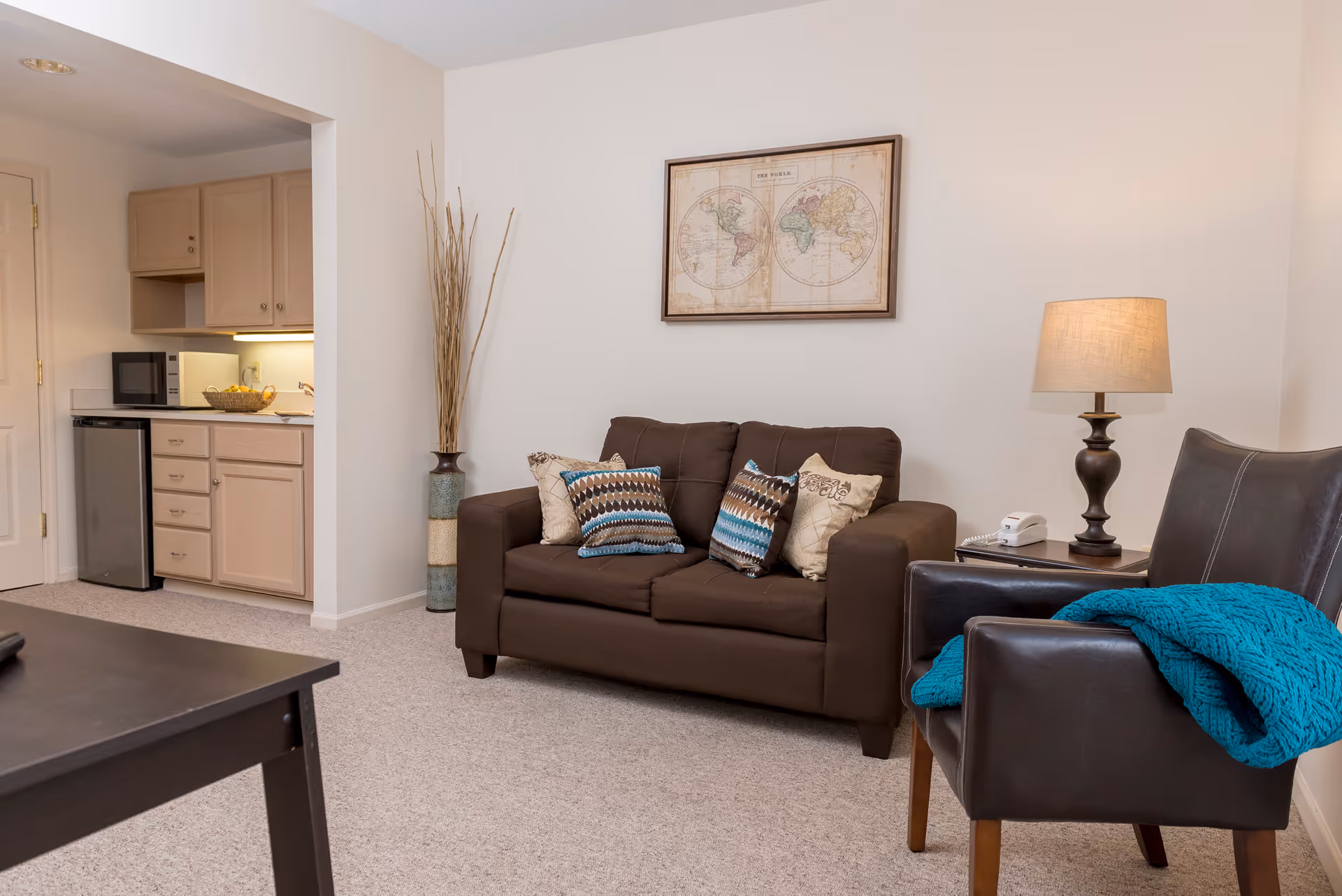 A cozy living room area featuring a brown loveseat with decorative pillows, a dark brown leather armchair with a teal throw blanket, a side table with a lamp and a telephone, and a framed world map on the wall. To the left, there is a small kitchenette with light wood cabinets, a microwave, a mini fridge, and a basket of fruit on the counter.