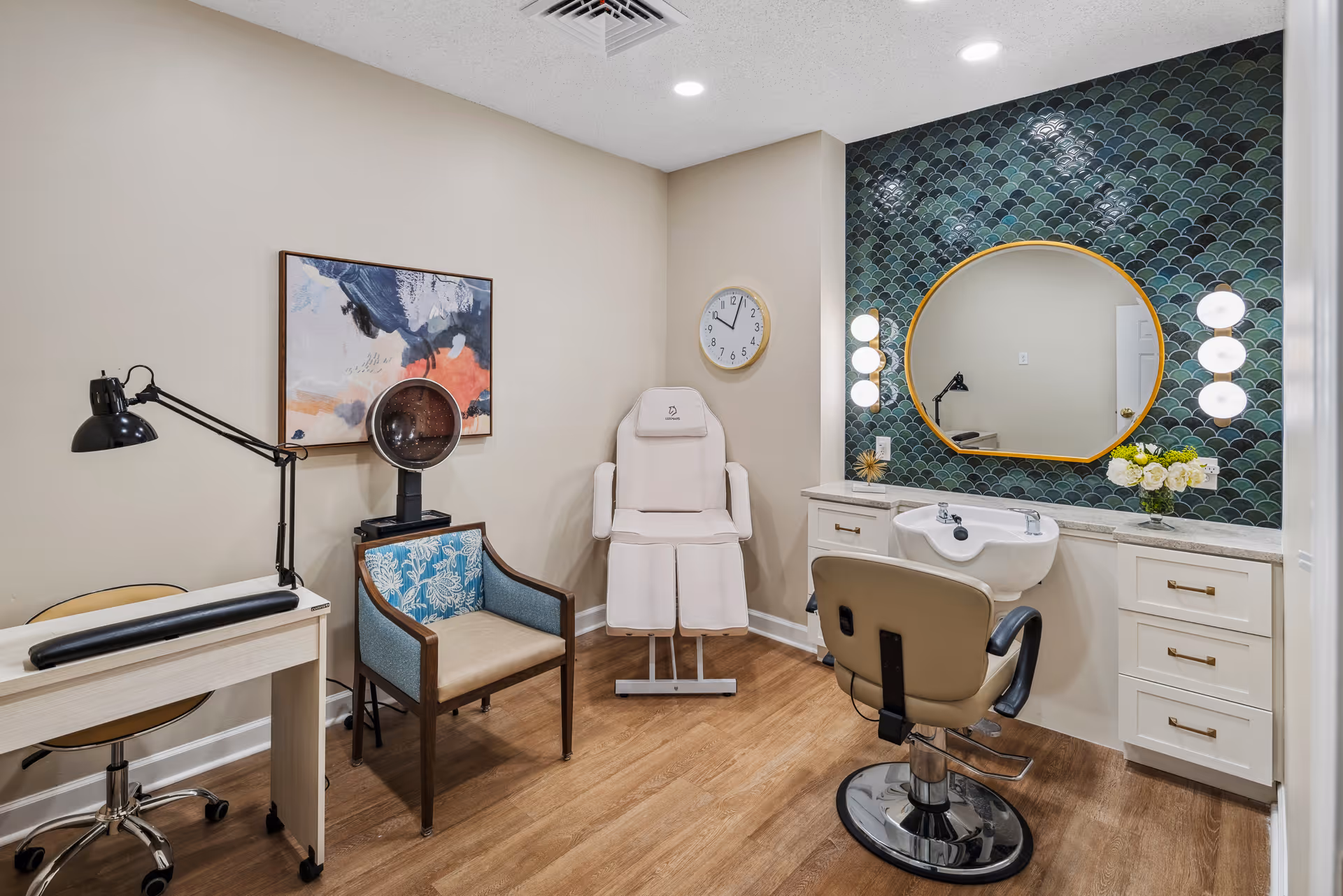 A well-lit beauty salon room with a salon chair in front of a sink and a large round mirror mounted on a wall with green fish scale tiles. To the left, there is a white manicure table with a black adjustable lamp and a blue cushioned chair with wooden arms. A white salon hair dryer chair and a wall clock are also visible. The floor is wooden and the walls are painted beige.