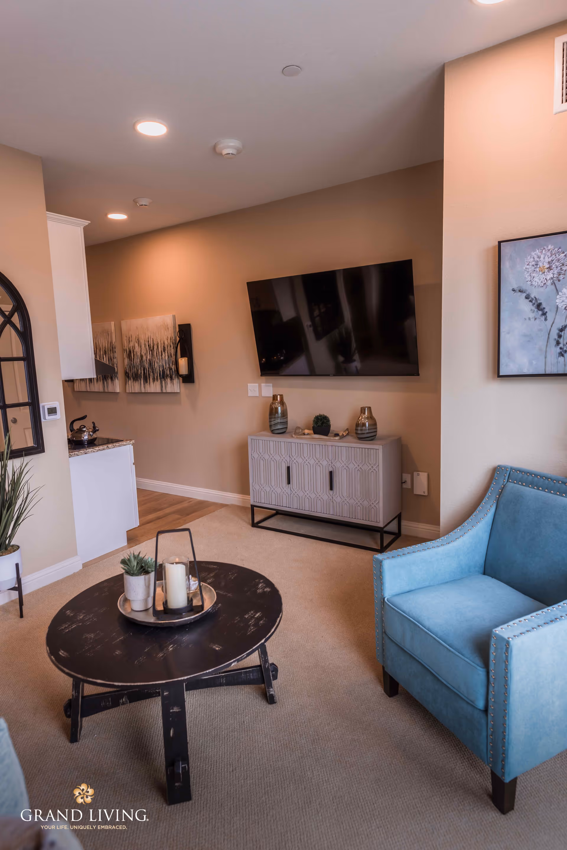A cozy living room area featuring a blue upholstered armchair with nailhead trim, a round black wooden coffee table with decorative items including a candle and small plants, a wall-mounted flat-screen TV above a patterned cabinet, and neutral beige walls adorned with framed artwork. The room has recessed ceiling lights and a glimpse of a kitchen area with granite countertops and a kettle.