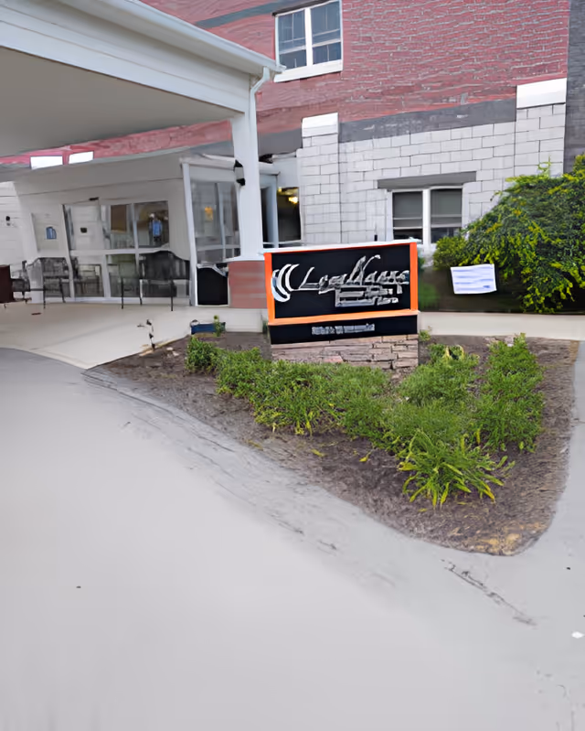 Entrance to Loyalhanna Senior Suites and Personal Care facility showing a covered drop-off area, a sign with the facility name, some greenery, and part of the building exterior with windows.