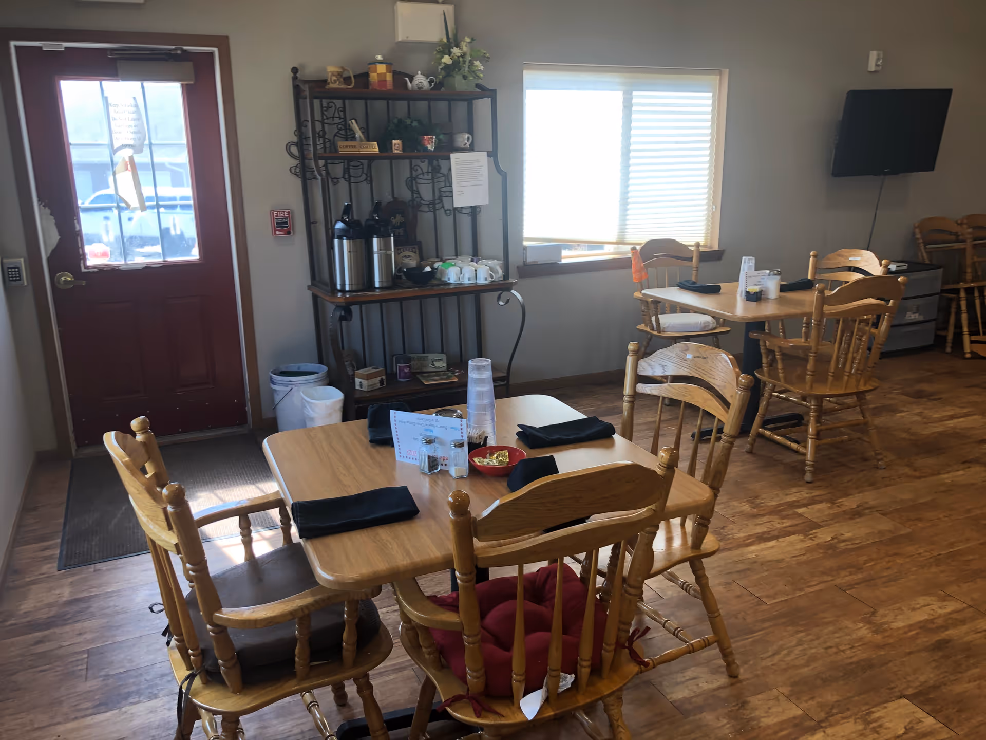 A dining area with wooden tables and chairs, each table set with black napkins and menus. There is a red door with a window on the left side, a window with blinds on the back wall, and a black metal shelf holding coffee dispensers, cups, and decorative items. A flat-screen TV is mounted on the right wall.
