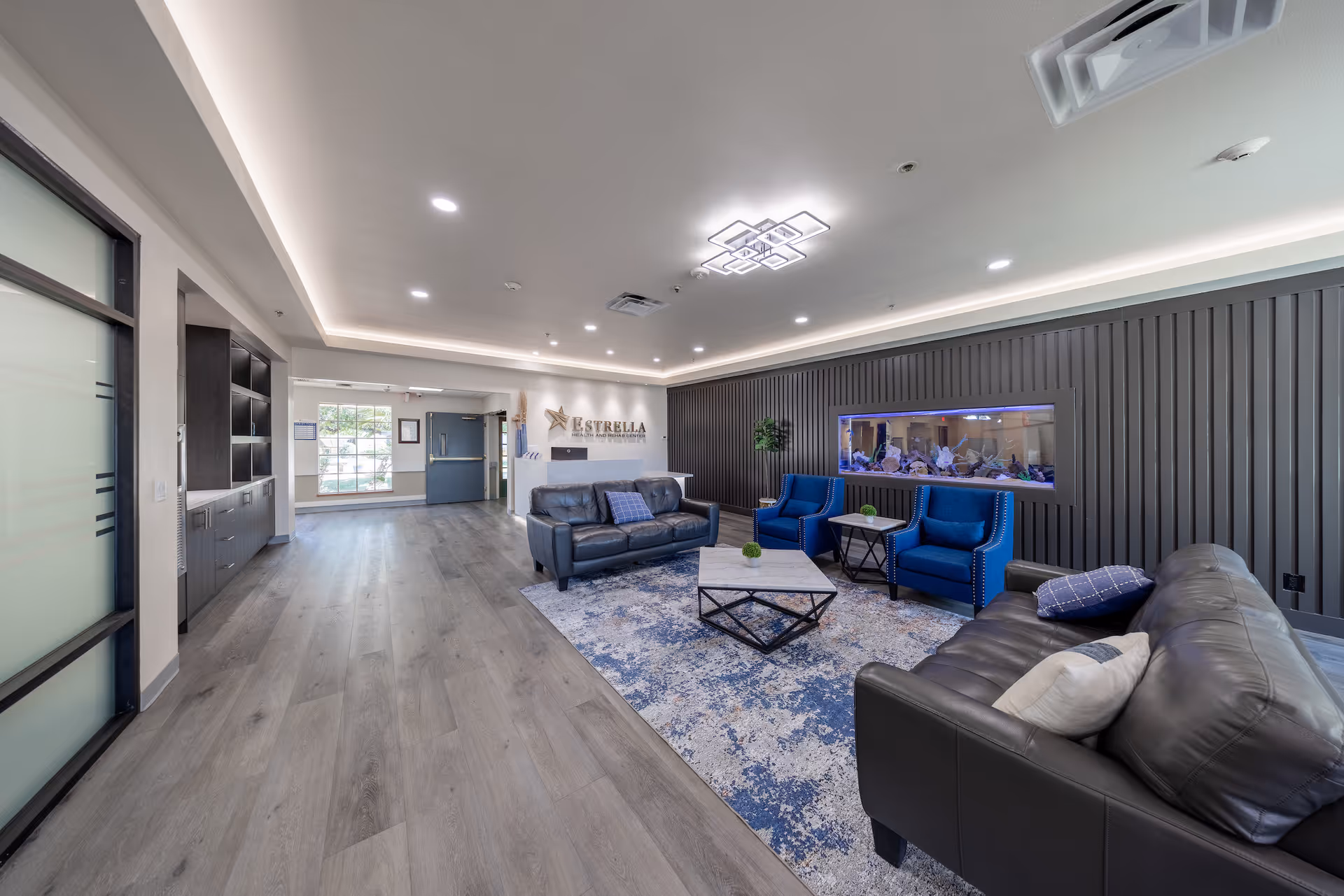 A modern and spacious lounge area inside Estrella Health and Rehab Center featuring gray leather sofas, two blue armchairs, a patterned area rug, a rectangular coffee table, and a built-in aquarium on a dark paneled wall. The room has recessed lighting, a decorative ceiling light fixture, and wood-look flooring. A hallway and large window are visible in the background.