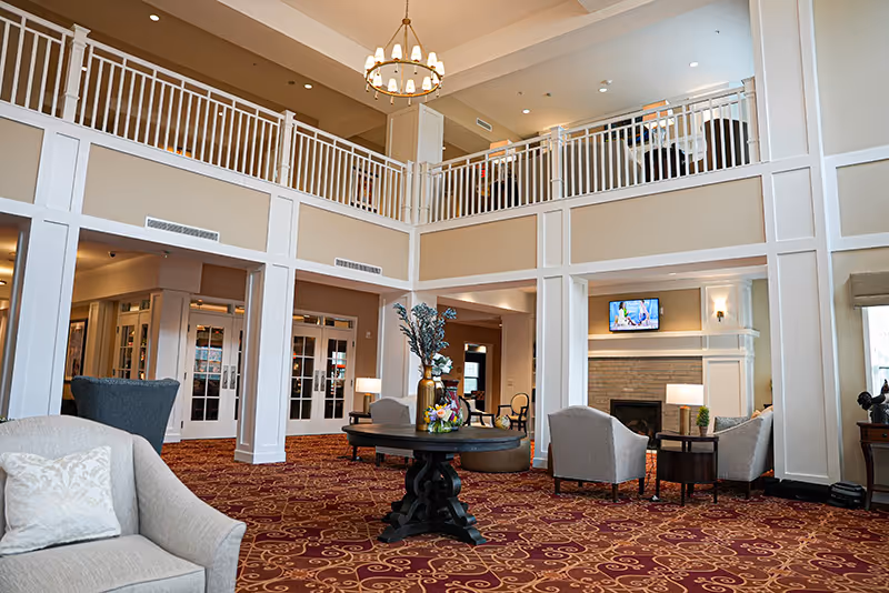 Spacious two-story lounge with seating, a central table and chandelier, and a fireplace in a senior living facility.