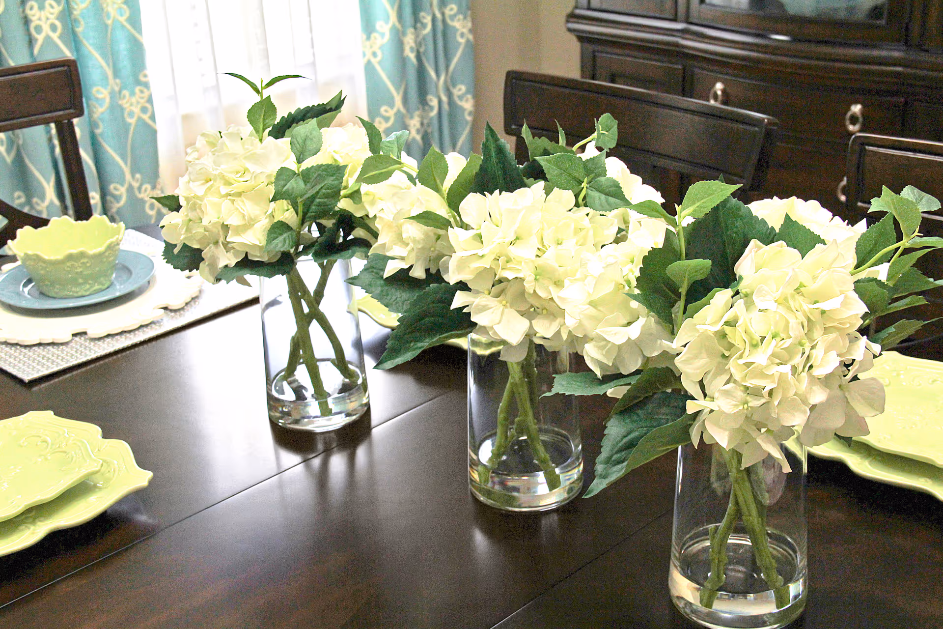 A dark wooden dining table set with three clear glass vases each holding a bouquet of white hydrangeas with green leaves. The table is also set with light green plates and bowls on placemats, with a background of blue curtains with a white pattern and dark wooden chairs.