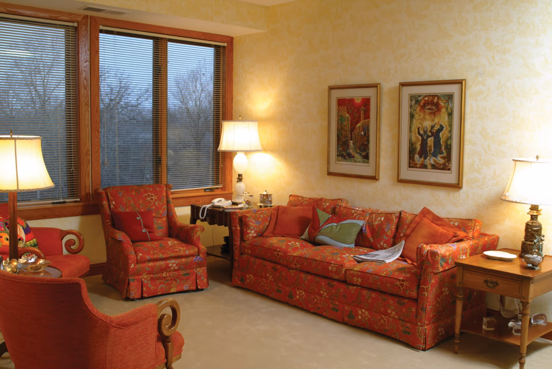 A cozy living room with a red floral-patterned sofa and matching armchairs. The room has beige walls with two framed paintings hanging above the sofa. There are three table lamps providing warm lighting, a wooden side table with a telephone, and a large window with closed blinds showing leafless trees outside.