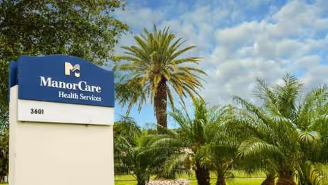 ManorCare Health Services sign with palm trees under a blue sky.