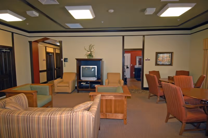 A cozy living room area in a retirement center featuring a striped sofa, two armchairs, a TV on a wooden stand, and a dining table with six upholstered chairs. The room has beige walls with dark trim, carpeted floor, and ceiling lights. A hallway and doorways are visible in the background.