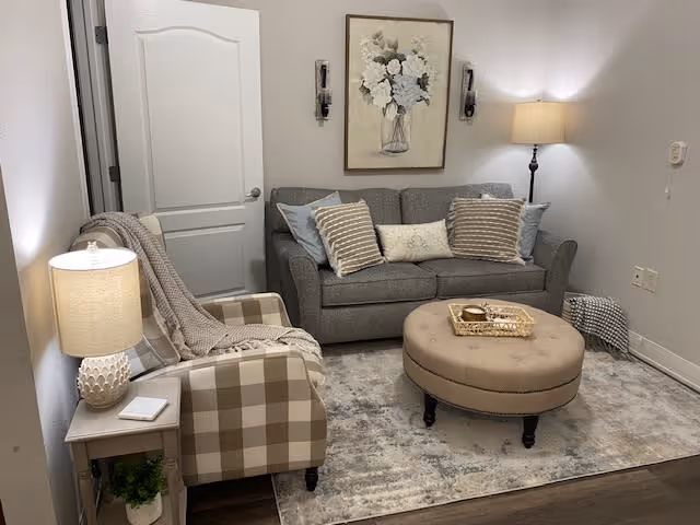 A cozy living room with a gray sofa adorned with multiple pillows, a round beige ottoman with a tray on top, a checkered armchair with a throw blanket, a side table with a lamp and a small plant, a floor lamp in the corner, and a floral painting on the wall above the sofa.
