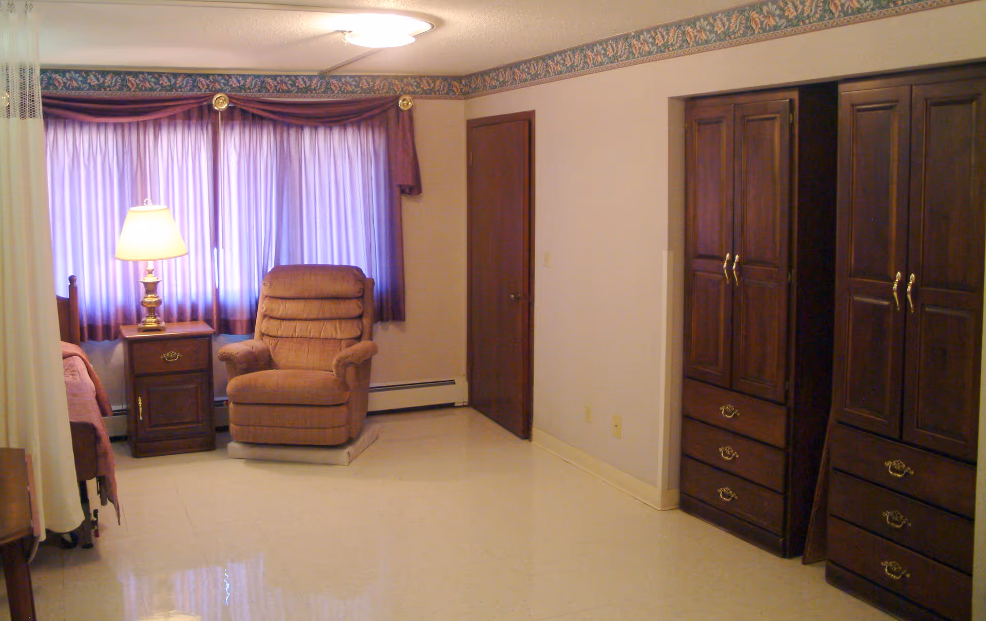 A room with a large window covered by purple curtains and a matching valance. In front of the window is a brown upholstered recliner chair and a wooden nightstand with a lamp. To the right, there are two large wooden wardrobes with brass handles and drawers. The floor is light-colored and shiny, and the walls are light with a floral wallpaper border near the ceiling.