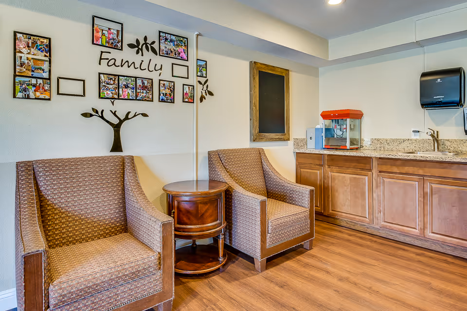 A cozy seating area in a senior living facility with two patterned armchairs and a round wooden side table between them. On the wall above the chairs is a decorative family photo collage with the word 'Family' and a tree design. To the right, there is a countertop with wooden cabinets, a popcorn machine, a sink, and a paper towel dispenser mounted on the wall.