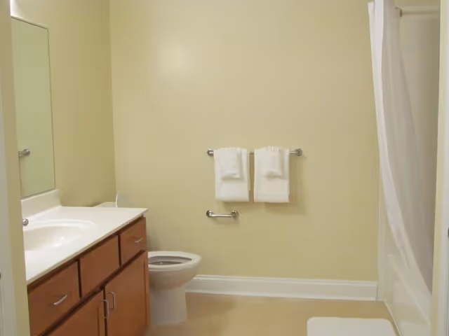 A clean bathroom with a white toilet, a wooden vanity with a white countertop and sink, a large mirror above the sink, two white towels hanging on a towel rack, and a bathtub with a white shower curtain.