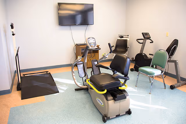 A small exercise room with various fitness equipment including a recumbent exercise bike, a treadmill, a stationary bike, and a green chair. A flat-screen TV is mounted on the wall above a small wooden cabinet.