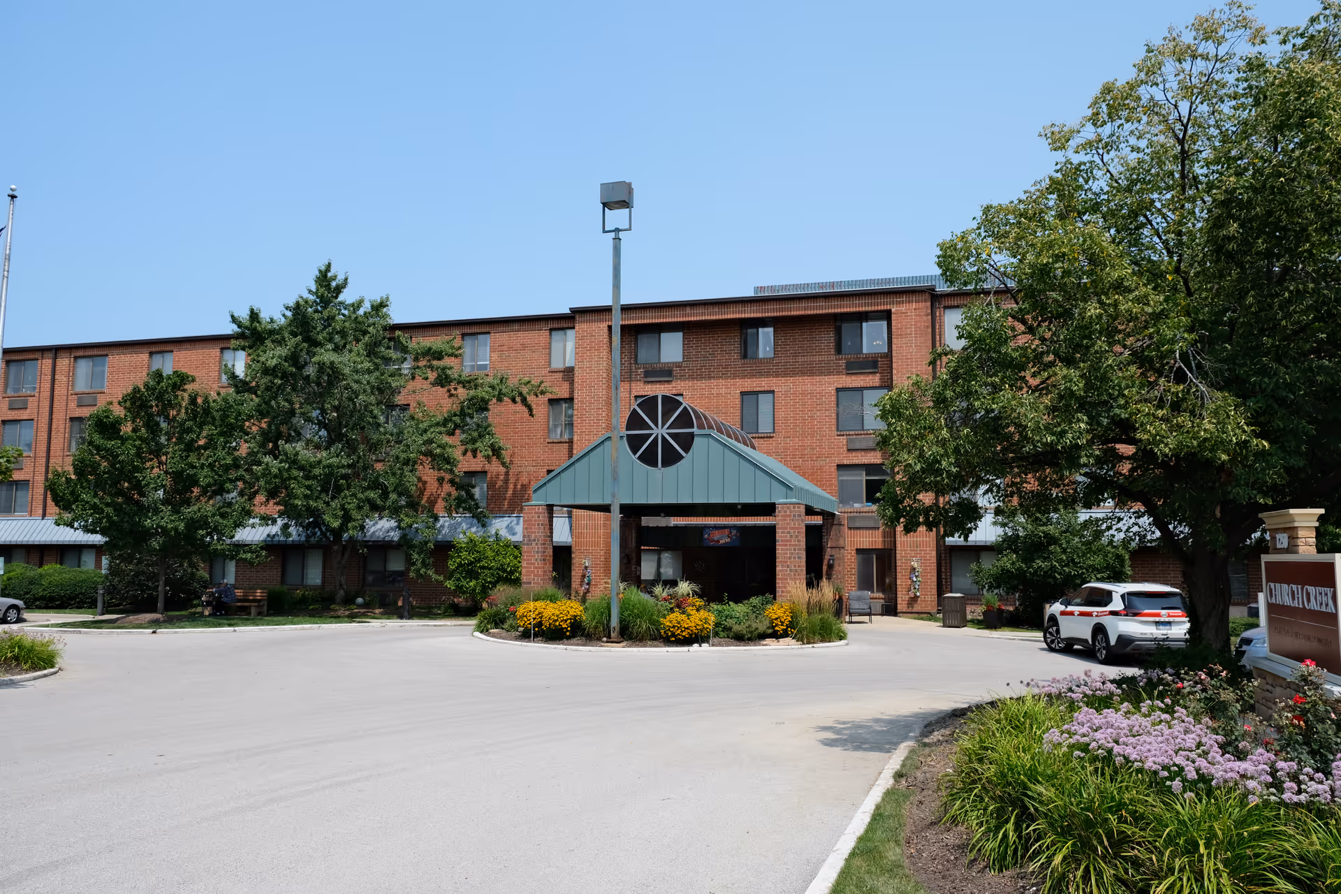 Front exterior view of Church Creek facility showing a multi-story brick building with a covered entrance, surrounded by trees, flowers, and parked cars under a clear blue sky.
