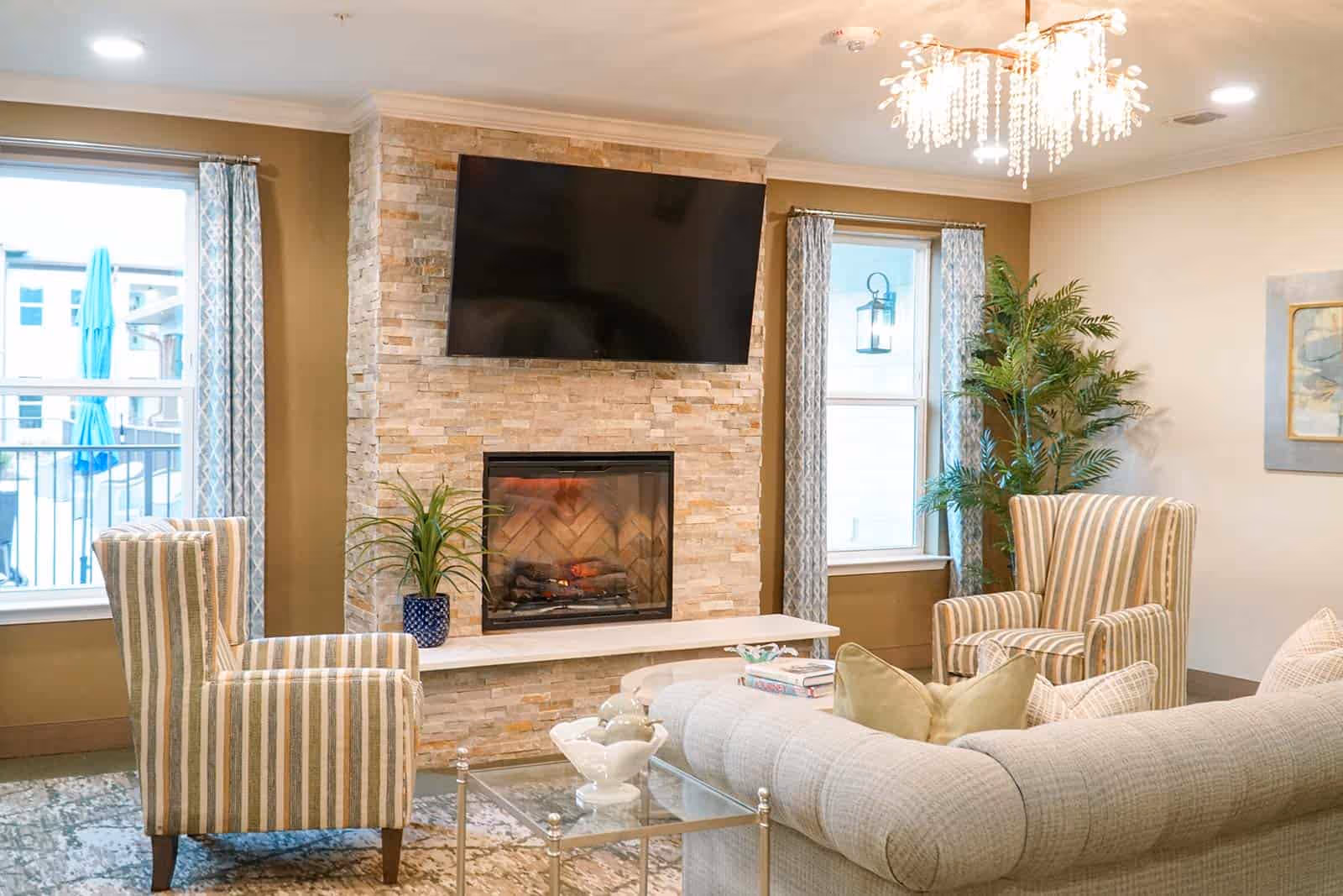A cozy living room with a stone fireplace and a mounted flat-screen TV above it. The room features two striped armchairs, a light-colored sofa with pillows, a glass coffee table with decorative items, two windows with patterned curtains, a potted plant, and a modern chandelier hanging from the ceiling.