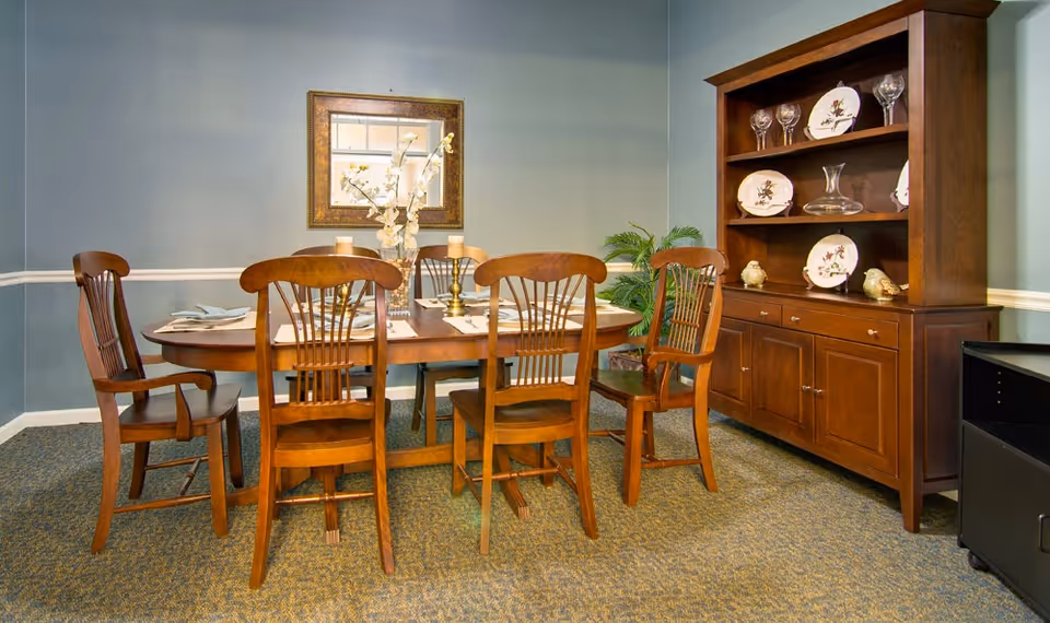 A dining room with a wooden dining table set for four, surrounded by five wooden chairs. On the table are placemats, napkins, and a centerpiece with white flowers and candles. Against the wall is a wooden hutch displaying decorative plates, glassware, and a decanter. A framed mirror hangs on the wall above the table, and a green plant is placed in the corner.