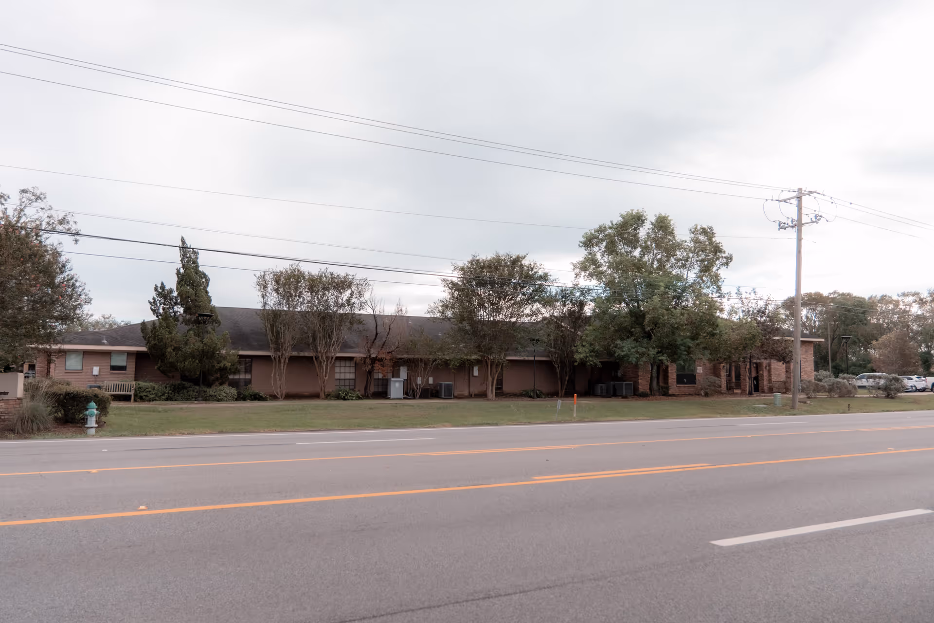 Single-story brick senior living building with trees and a lawn facing a road under a cloudy sky.