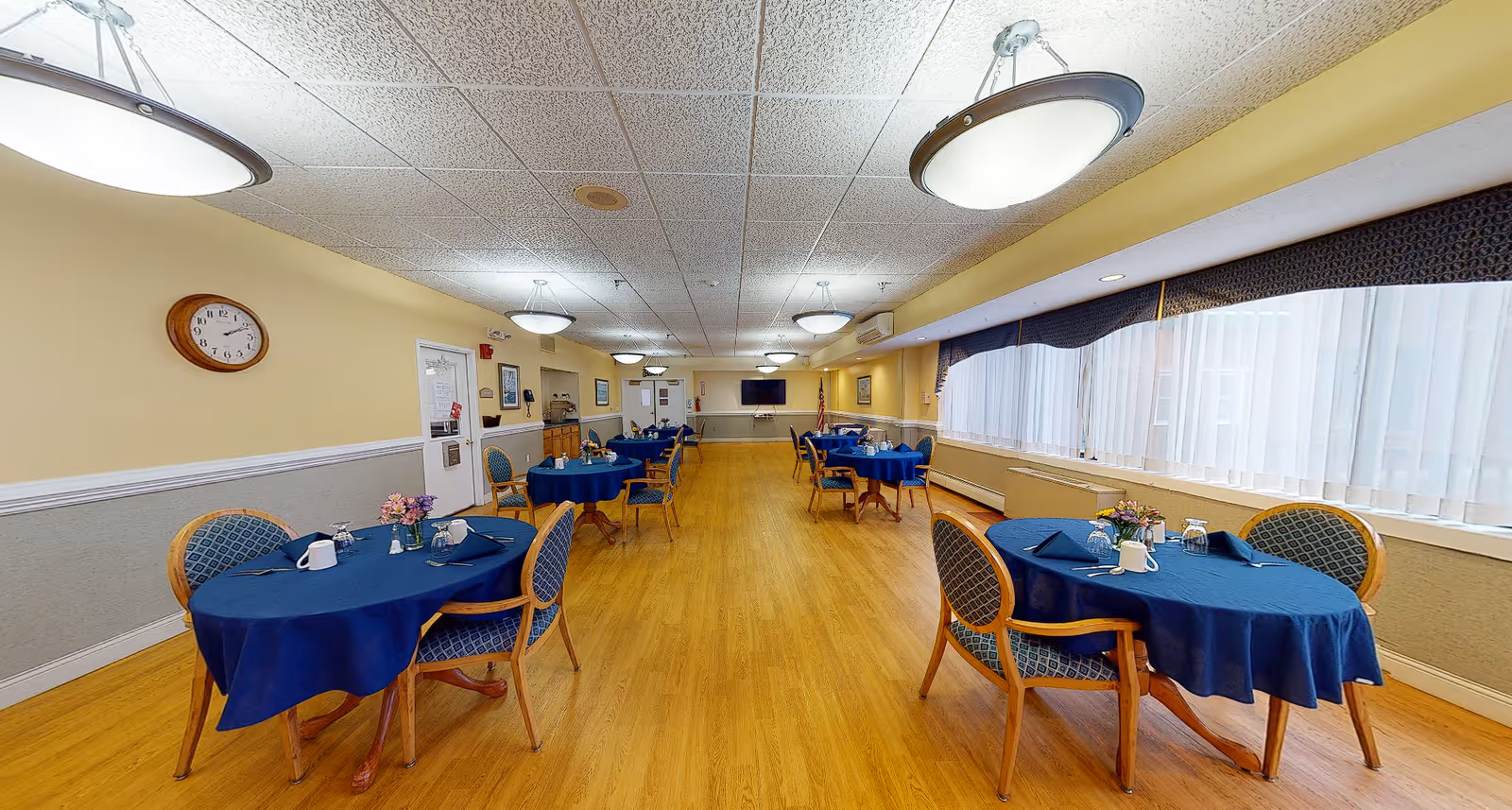 Large dining room with round tables draped in blue tablecloths, wooden chairs, hardwood floor and windows along the right wall.