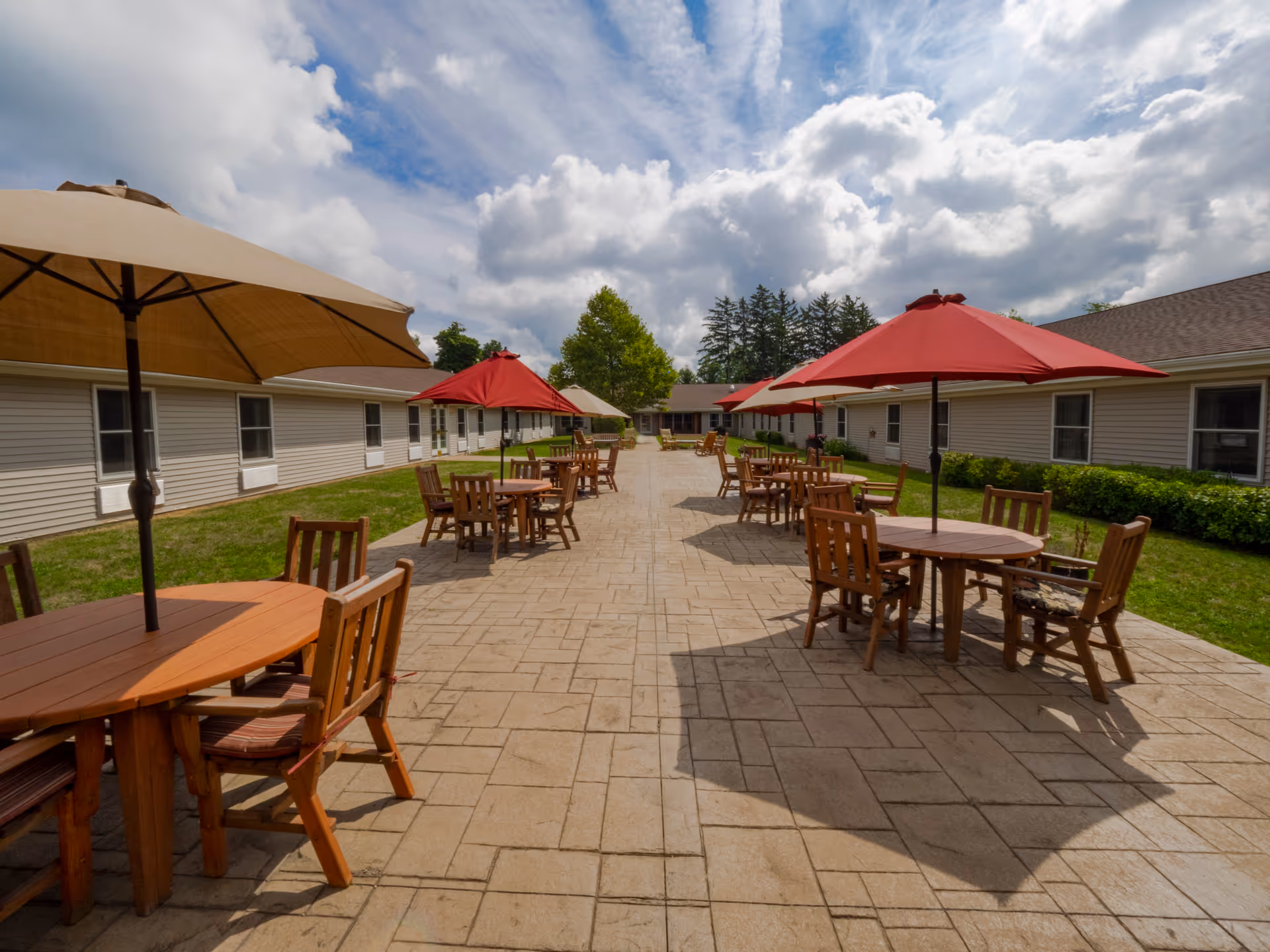 Outdoor courtyard with multiple wooden patio tables and umbrellas between single-story buildings under a partly cloudy sky.