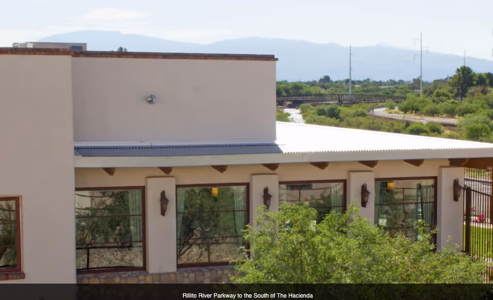 Exterior view of a single-story building with large windows and a metal roof, surrounded by greenery and trees, with mountains visible in the background under a clear sky.