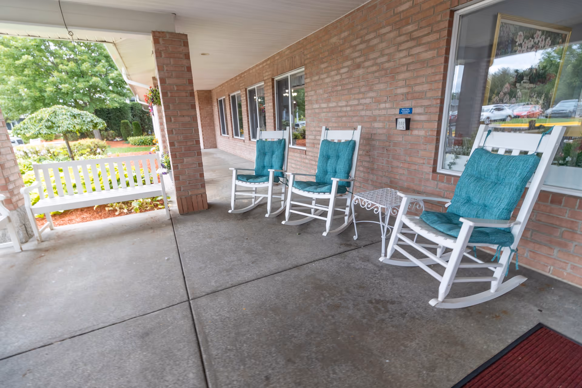 Covered outdoor patio area with three white rocking chairs with teal cushions and a small white metal table between two of the chairs. A white bench is visible on the left side near a brick column. The patio overlooks a garden with green trees and shrubs.