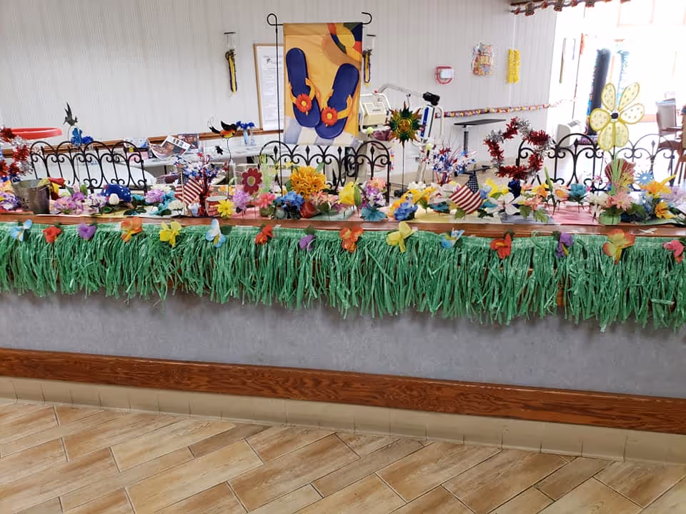 A decorated counter with a green grass skirt and colorful artificial flowers, butterflies, and small American flags. Behind the counter are black wrought iron chairs and a wall with a hanging decoration featuring blue flip-flops with orange flowers. The setting appears to be a common area or reception space inside a facility.