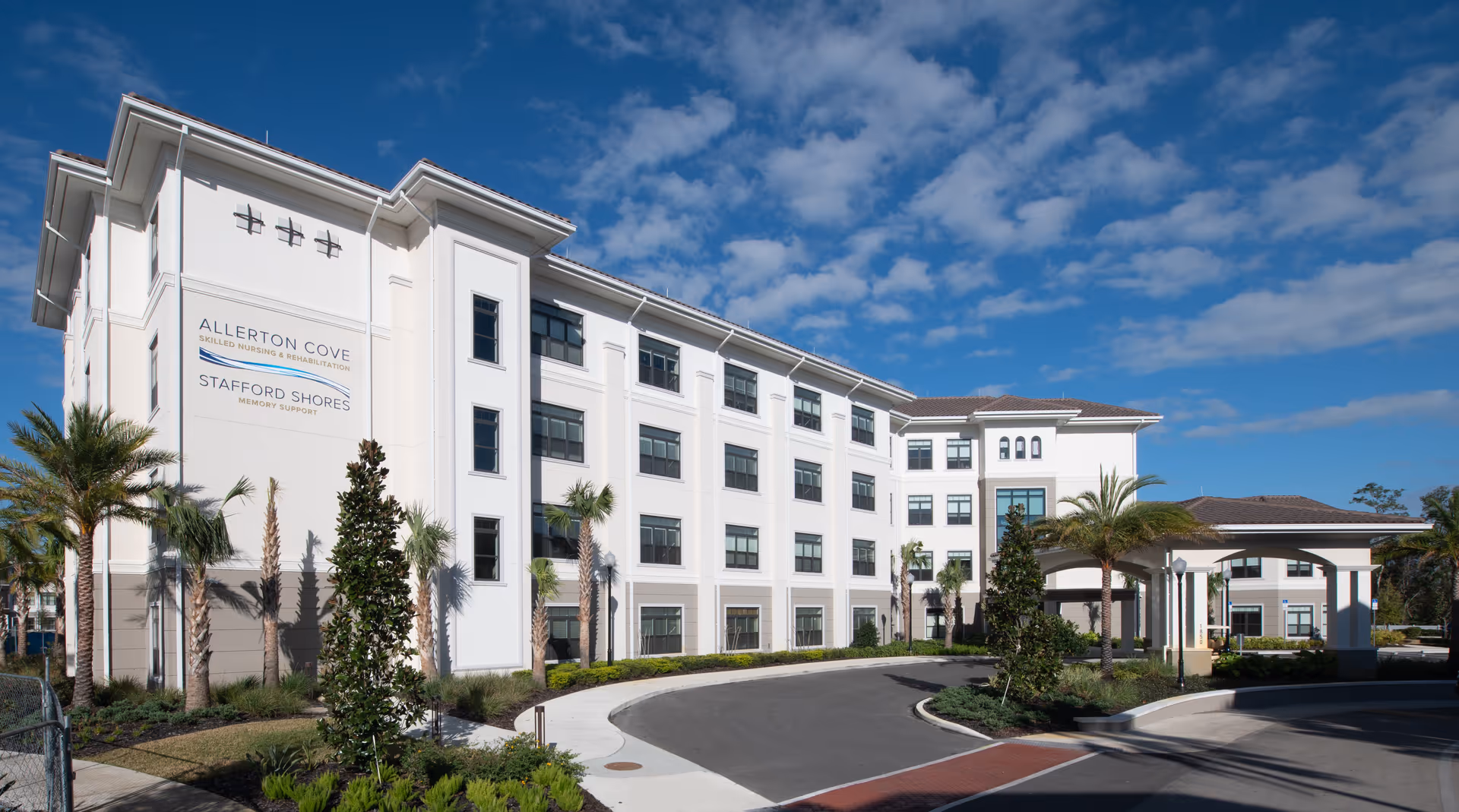Exterior view of a large, modern, white senior living facility building with multiple windows, palm trees, and a curved driveway under a partly cloudy blue sky.