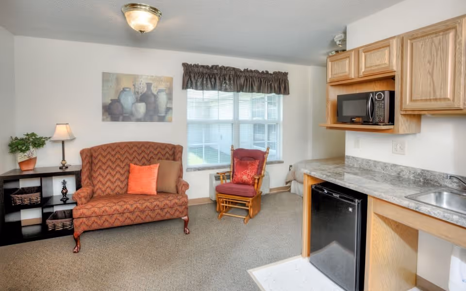 A cozy living area in a senior living facility featuring a patterned loveseat with two cushions, a wooden rocking chair with a red cushion, a small black shelving unit with baskets and a lamp, a window with blinds and a valance, and a kitchenette with wooden cabinets, a microwave, a small refrigerator, and a sink.