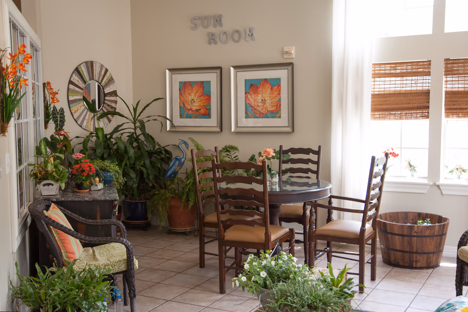 A bright sunroom with a round glass-top table surrounded by four wooden chairs with tan cushions. The room is decorated with various green plants and flowers in pots, a wicker chair with a striped cushion, and two framed floral paintings on the wall. The words 'SUN ROOM' are displayed above the paintings. Large windows with bamboo blinds allow natural light to fill the space.