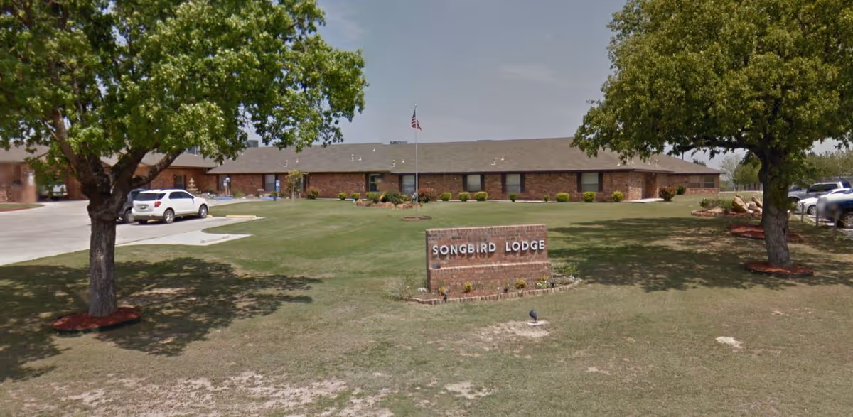 Exterior view of Songbird Lodge, a single-story brick building with a large grassy lawn, two trees, a flagpole, and a sign displaying the facility's name. Several cars are parked near the building under a clear sky.