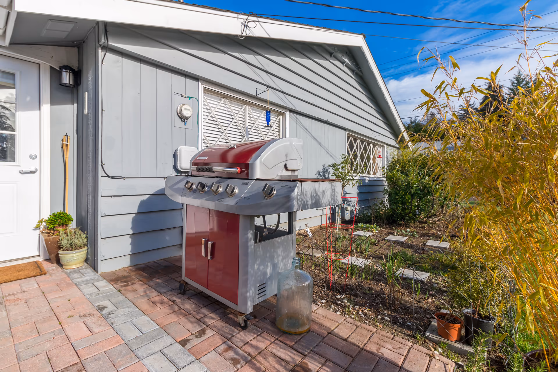 Outdoor patio area with a red and silver gas grill on wheels placed on a brick-paved surface next to a light blue house with white-framed windows. There are some potted plants near the white door and garden plants growing in the soil beside the patio. The sky is clear with some clouds and power lines are visible overhead.