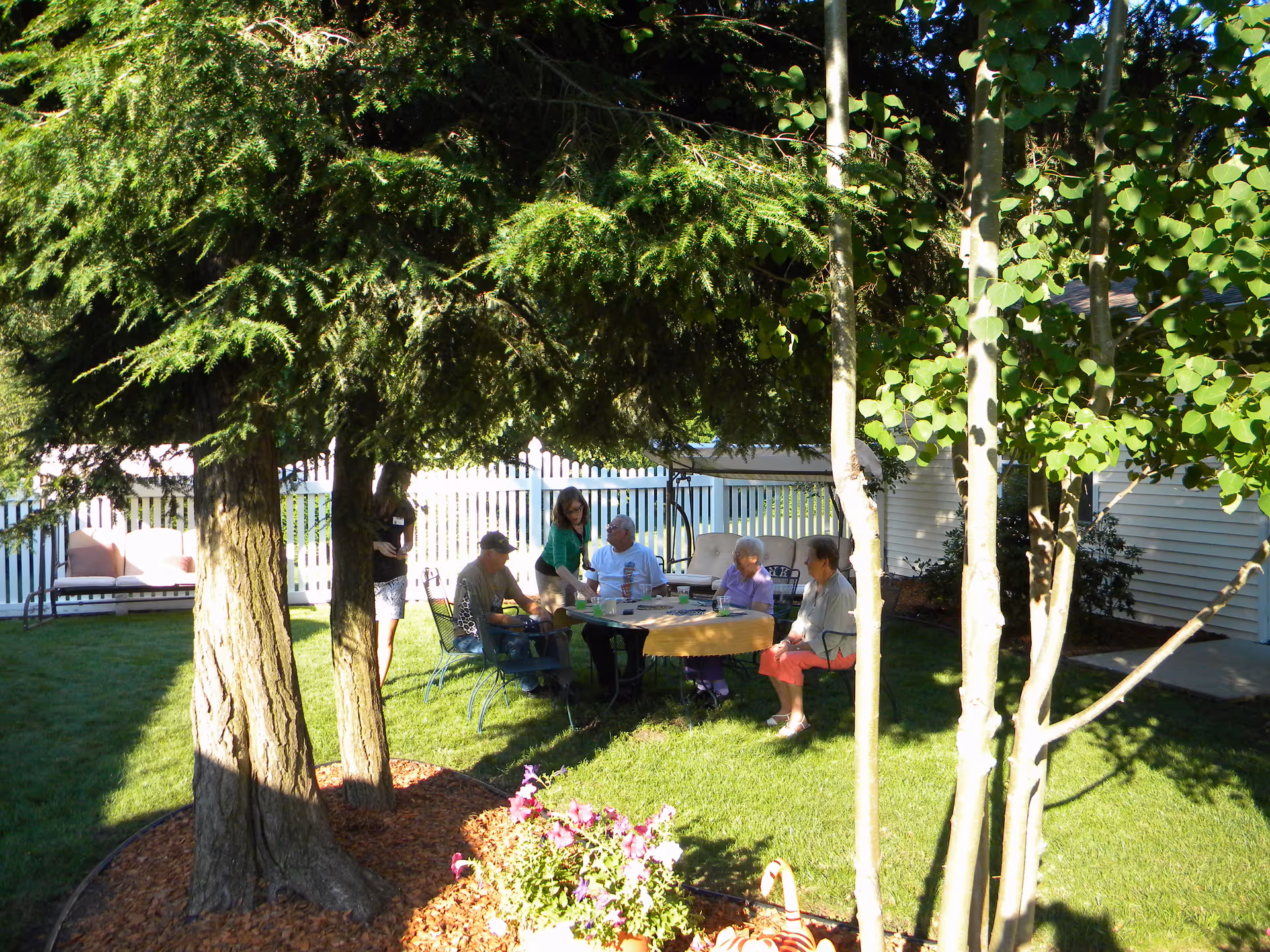 A group of people seated around an outdoor table under trees in a fenced backyard garden.