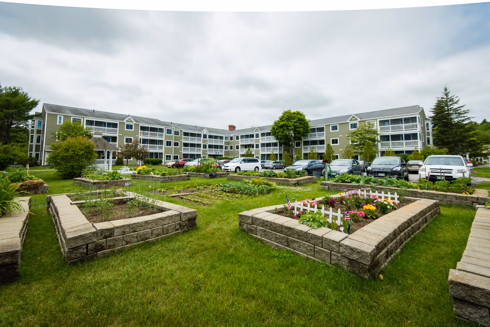 Exterior view of a multi-story senior living building with raised garden beds, lawn, and parked cars in front.