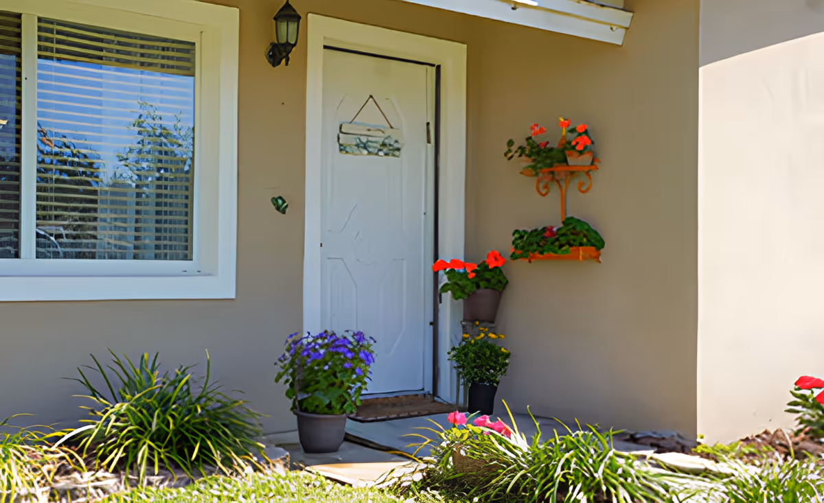 Front entrance of a light-colored house with a white door, window, porch light and several potted and wall-mounted flowering plants.