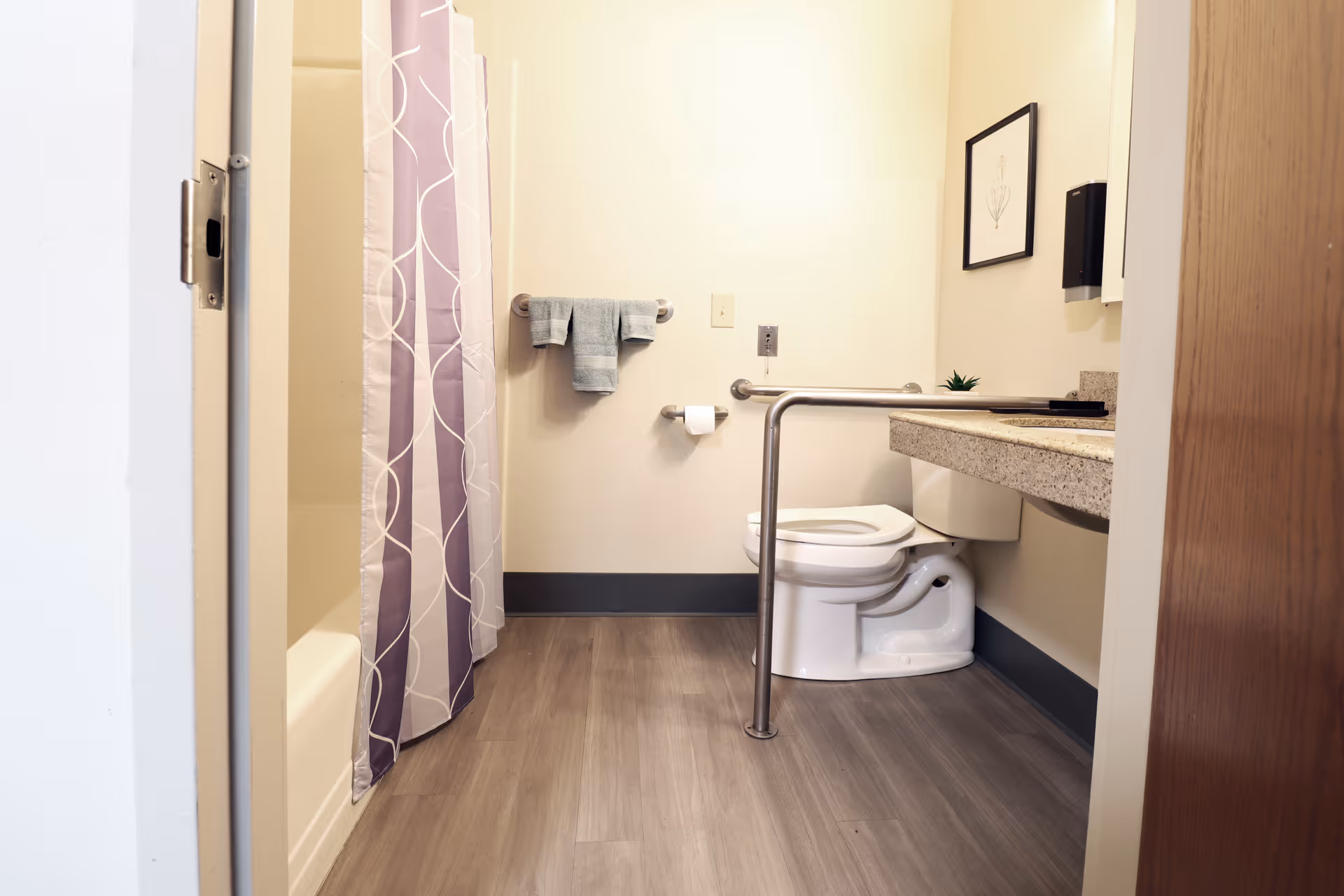 A clean and accessible bathroom with a toilet equipped with a safety grab bar, a countertop with a sink, a towel rack with a folded towel, a toilet paper holder, and a shower with a purple and white patterned curtain.