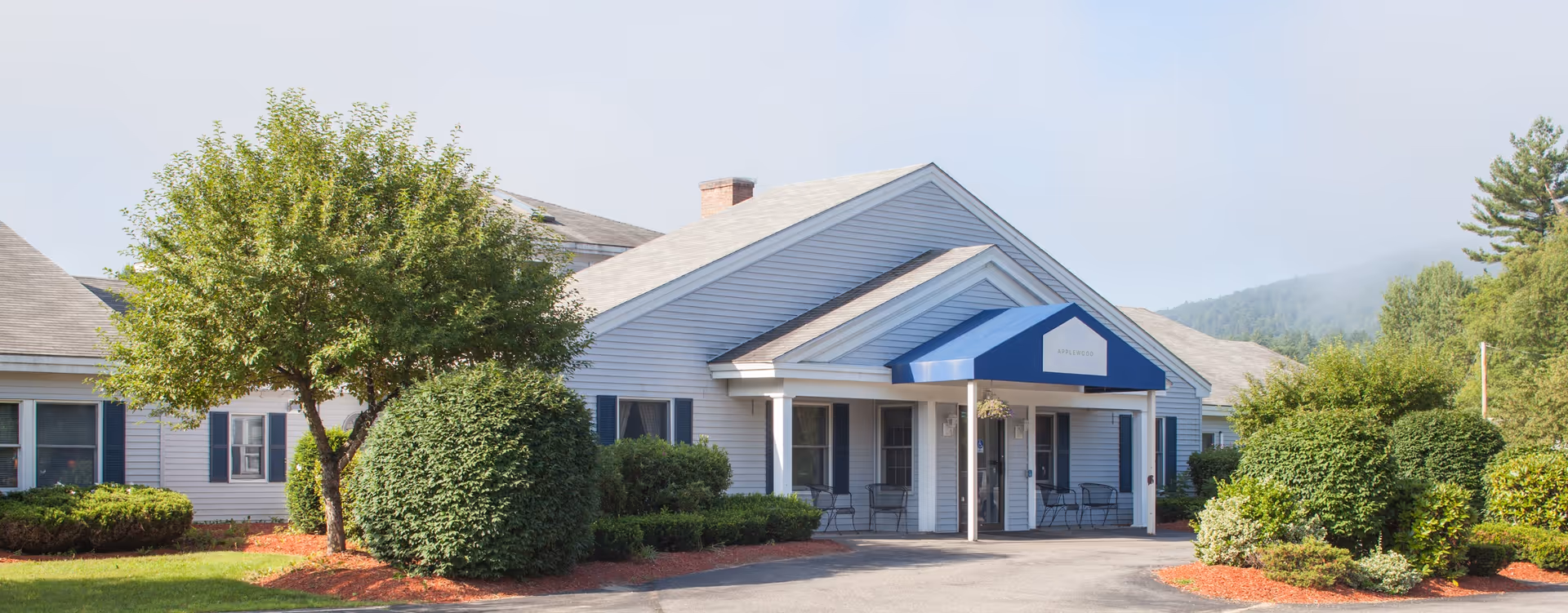 Exterior view of a single-story building with white siding and a blue awning over the entrance. The building is surrounded by well-maintained bushes, trees, and landscaped areas with red mulch. The sky is clear with some light clouds and there are hills visible in the background.