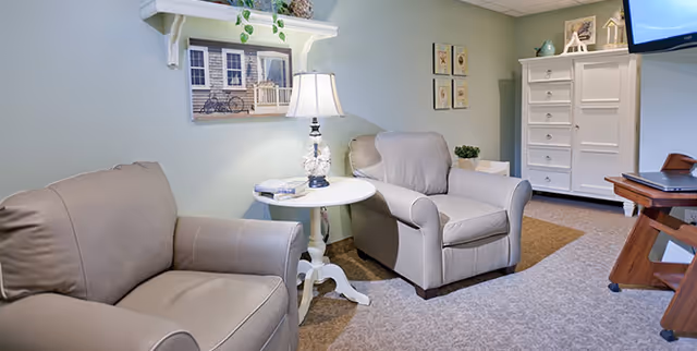 A cozy living room area with two beige armchairs, a small white round table with a lamp and books, a white cabinet, and a wall-mounted TV. The walls are painted light green and decorated with framed pictures and a shelf with plants.