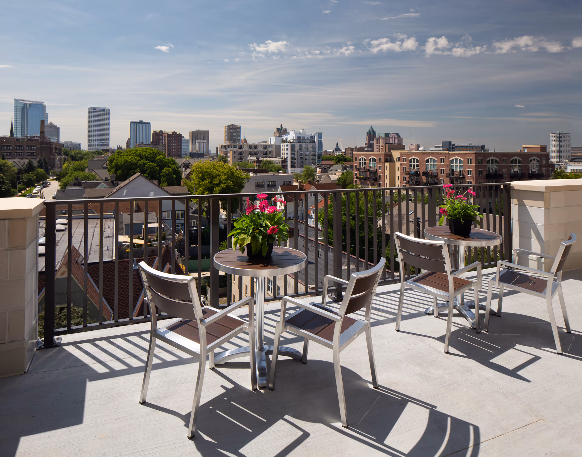 Outdoor balcony area with two small round tables, each with a potted plant with pink flowers, and four metal chairs. The balcony overlooks a cityscape with various buildings and trees under a partly cloudy sky.