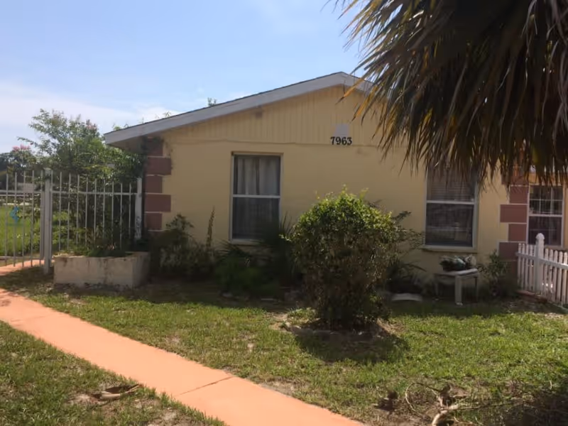 Exterior view of a single-story yellow building with two windows, a small garden with bushes, a white fence on the right, and a metal gate on the left. The building number 7963 is visible near the roof. The sky is clear and blue.