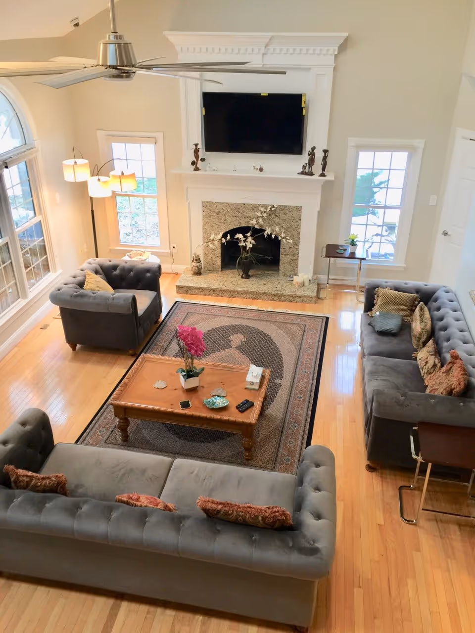 A bright living room with three gray tufted sofas arranged around a wooden coffee table on a patterned rug. The room features a white fireplace with a mounted flat-screen TV above it, decorative statues on the mantel, and a vase with flowers inside the fireplace. There are two windows on either side of the fireplace, a modern floor lamp with three lights, and a ceiling fan with metal blades. The floor is light wood, and the room has a cozy, inviting atmosphere.