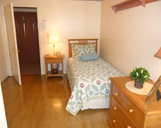 A small bedroom with a single bed against a pale wall, a nightstand with a lamp, a wooden dresser and an open door.