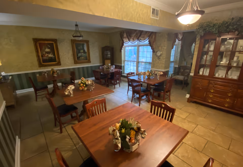 A cozy dining room with several wooden tables and chairs arranged neatly. Each table has a floral centerpiece and decorative napkins. The room features a large window with curtains, framed paintings on the walls, and a wooden china cabinet filled with white dishes. The floor is tiled and the lighting is warm and inviting.