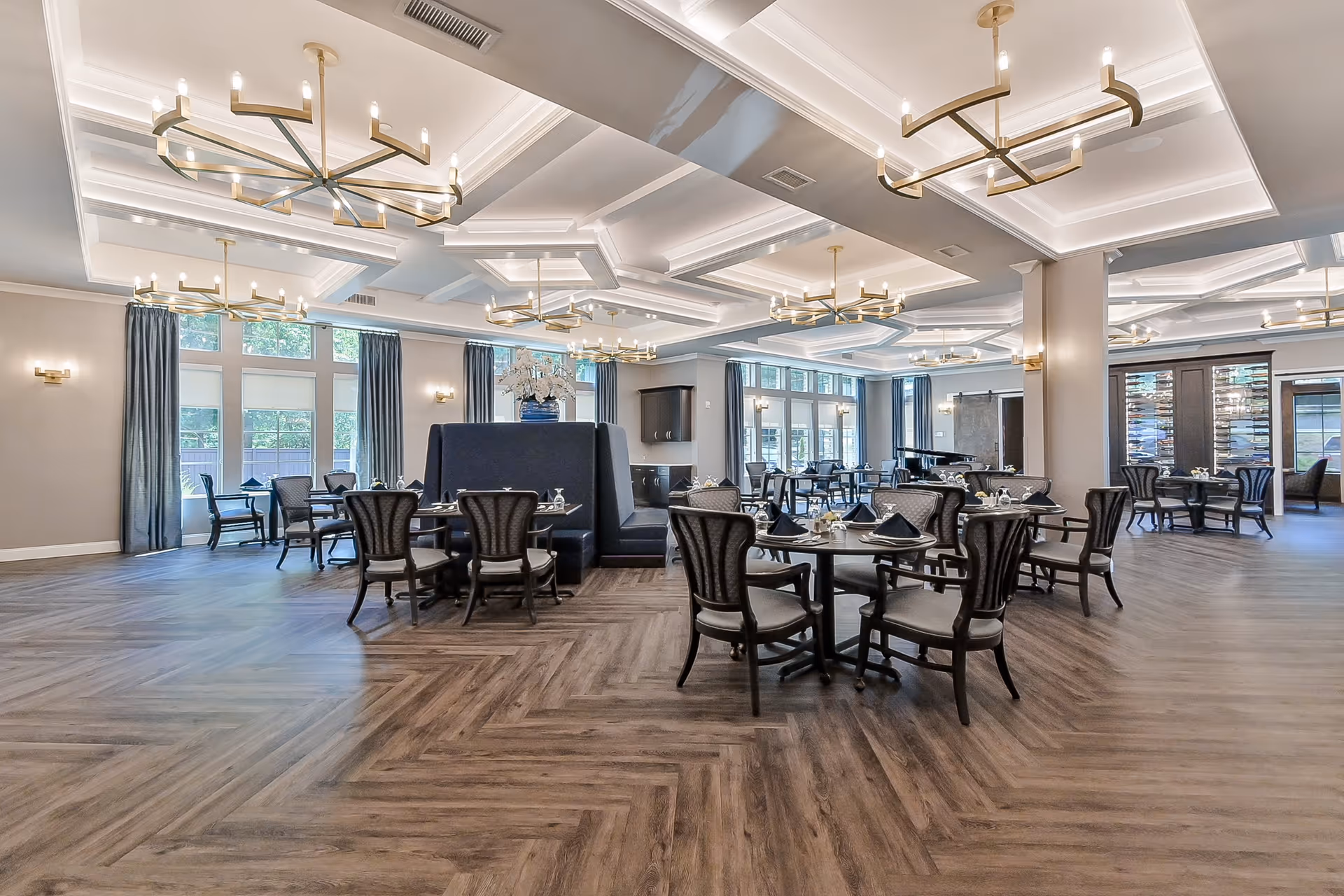 Spacious, elegant dining room with round tables and upholstered chairs under decorative chandeliers and large windows.