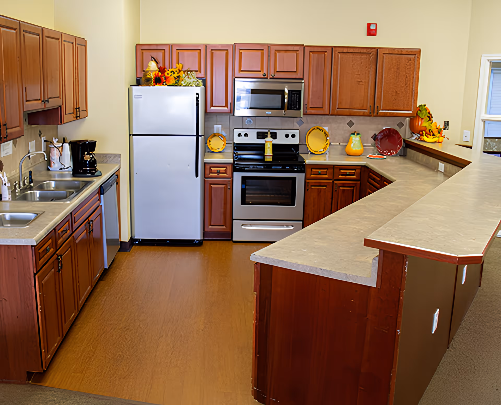 A clean and well-organized kitchen with wooden cabinets, a white refrigerator, a stainless steel oven with a microwave above it, a double sink, a dishwasher, and a long countertop with decorative plates and artificial fruit arrangements.