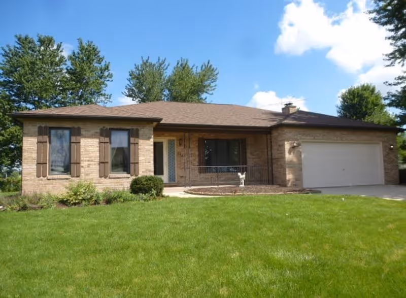 Single-story brick house with a brown roof, two windows with brown shutters on the left, a front door with glass panels, a small porch with a railing, and a two-car garage on the right. The house is surrounded by a green lawn and trees under a partly cloudy blue sky.