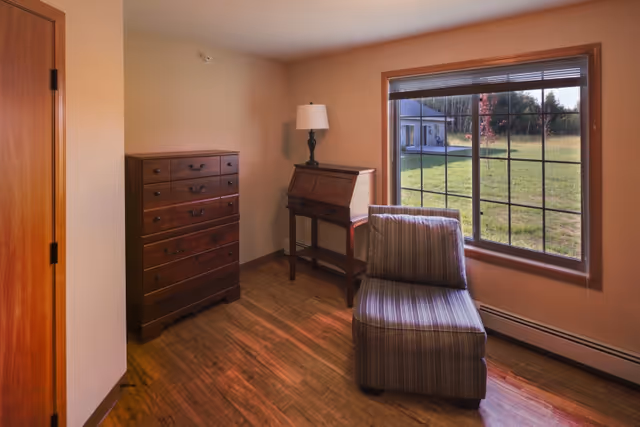 Small seating area with a striped upholstered chair, wooden chest of drawers and a writing desk with lamp beside a large window overlooking a grassy yard.