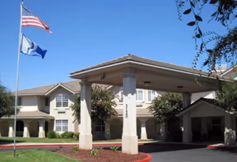 Front entrance of a two-story senior living building with a covered porte-cochere, flagpoles, and landscaped lawn.