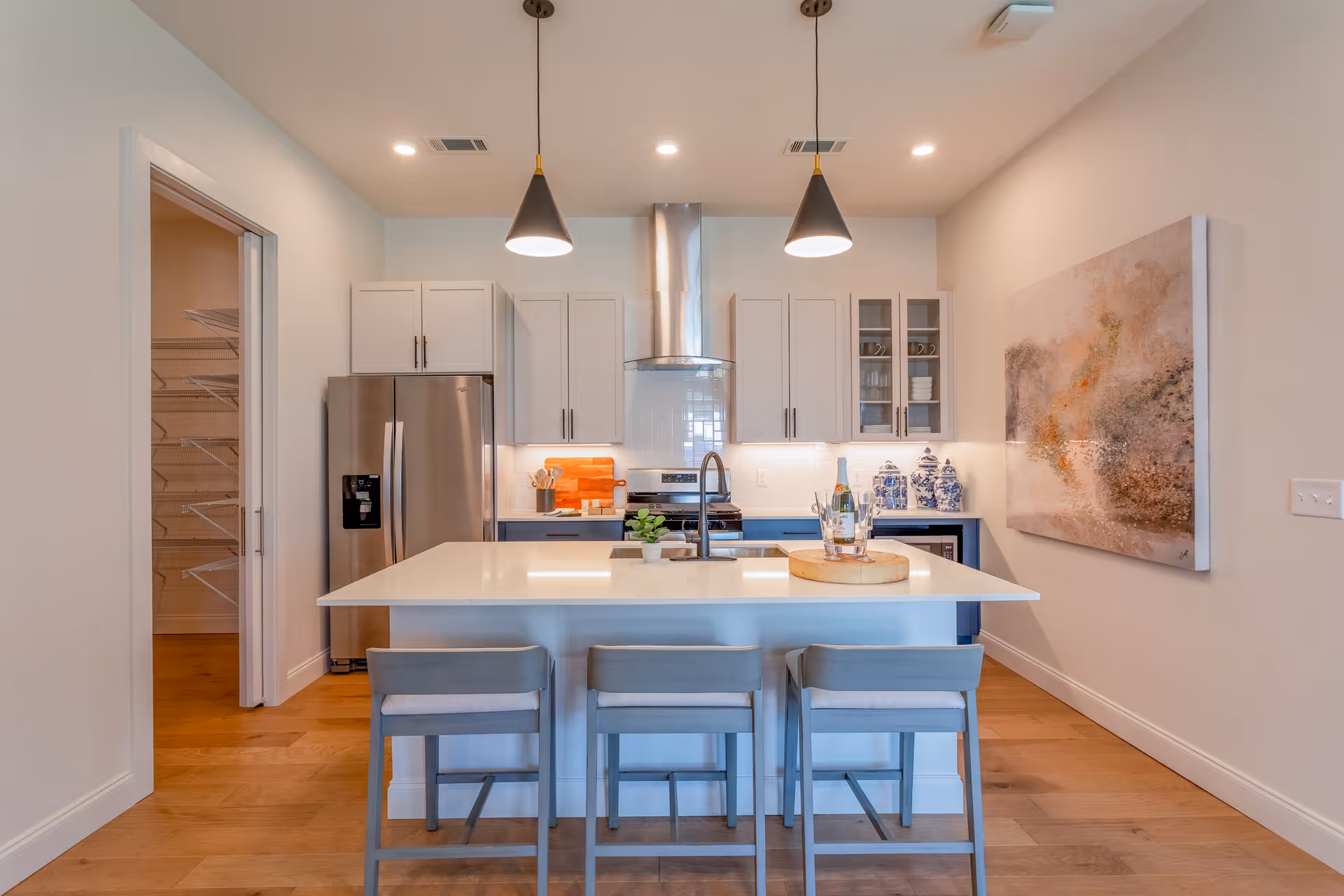 Modern kitchen with white cabinetry, stainless steel refrigerator, stove with a stainless steel hood, and a large white island with three gray bar stools. Two pendant lights hang above the island. There is a pantry with wire shelving visible through an open door on the left. A decorative tray with a bottle and glasses is on the island, and a large abstract painting is on the right wall.