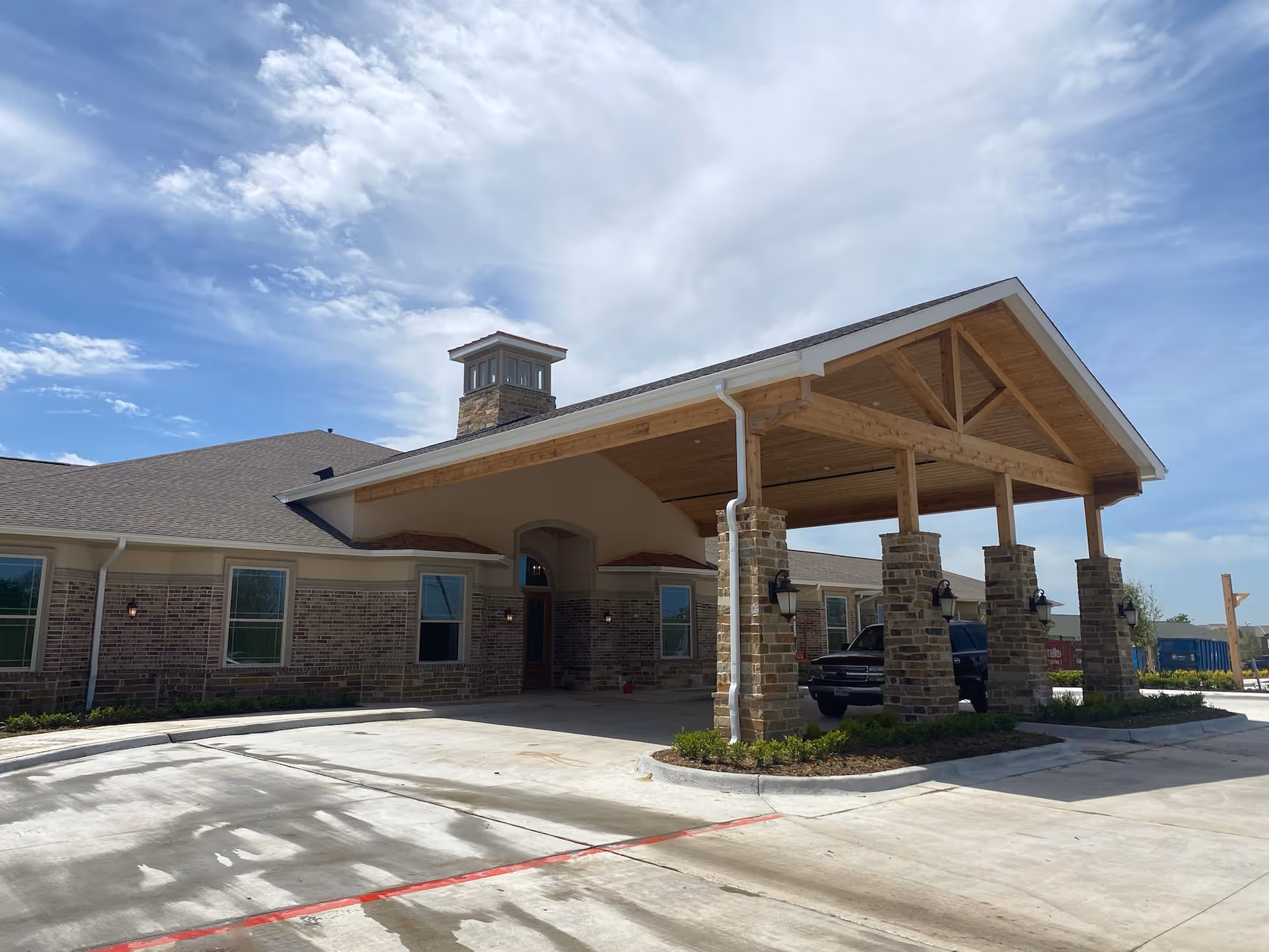 Front entrance of a single-story brick memory care building with a large covered porte-cochere supported by stone columns under a partly cloudy sky.